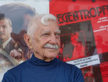 Porträt eines älteren Mannes (Harry Raymon) mit grauem Haar und Schnurrbart, im Hintergrund ist ein Filmplakat zu sehen.