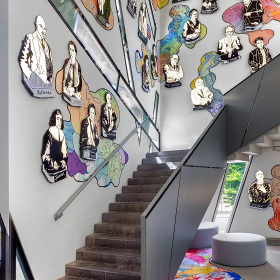 Bunte, comicartige Büsten berühmter Persönlichkeiten an den Wänden eines Treppenhauses, unten Sitzmöbel