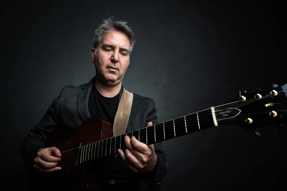 Ein Mann mit kurzen grauen Haaren im grauen Sacko spielt Gitarre und schaut dabei den Gitarrenhals an.