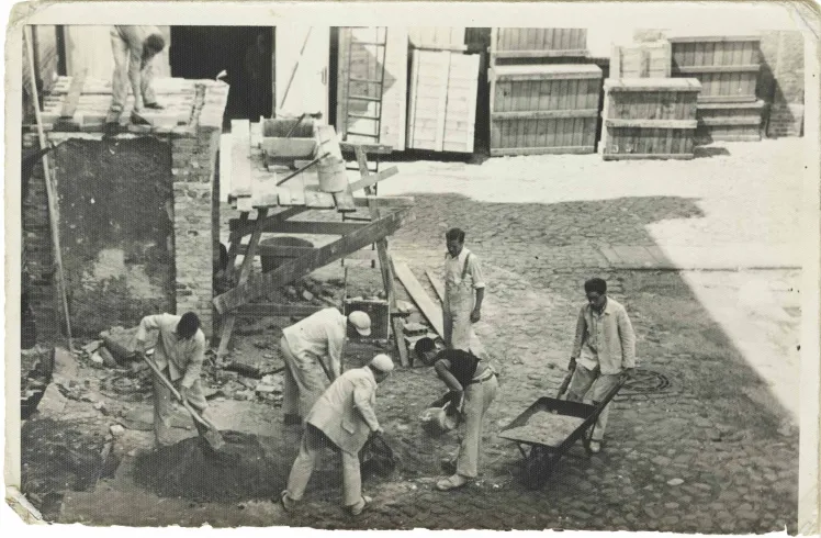 Sieben Männer in Arbeitskleidung und mit Schubkarre, Schaufeln und weiterem Arbeitsgerät (Schwarz-Weiß-Foto).